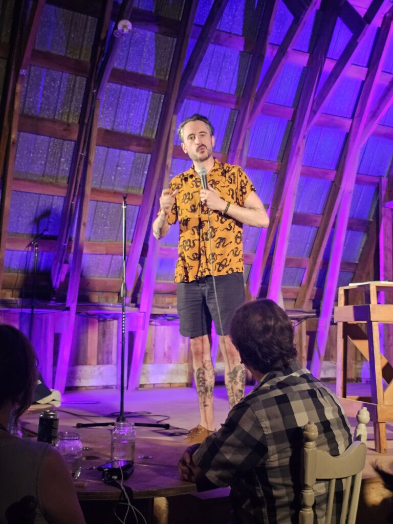 Joe Guérin en pleine performance sur la scène de La Ferme Kerr, vêtu d'une chemise jaune et éclairé par une lumière violette, capturant son énergie comique unique