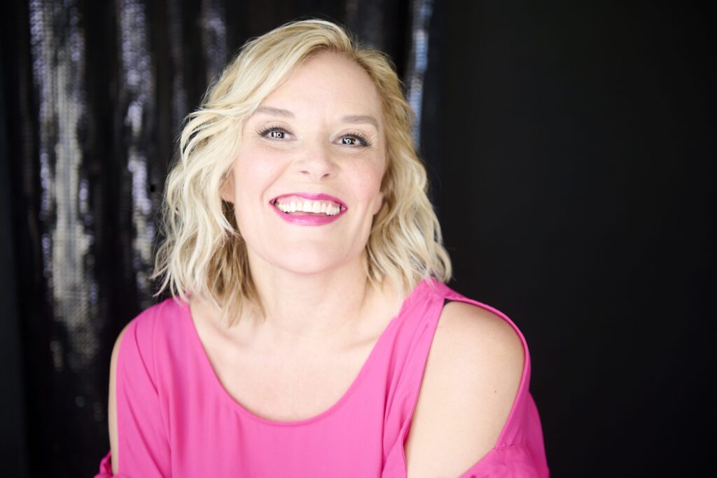Portrait lumineux de Rachelle Elie souriante, humoriste bilingue portant une tenue rose vif devant un arrière-plan sombre